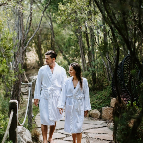man and woman in white bath robes walking along path in nature surrounded by trees