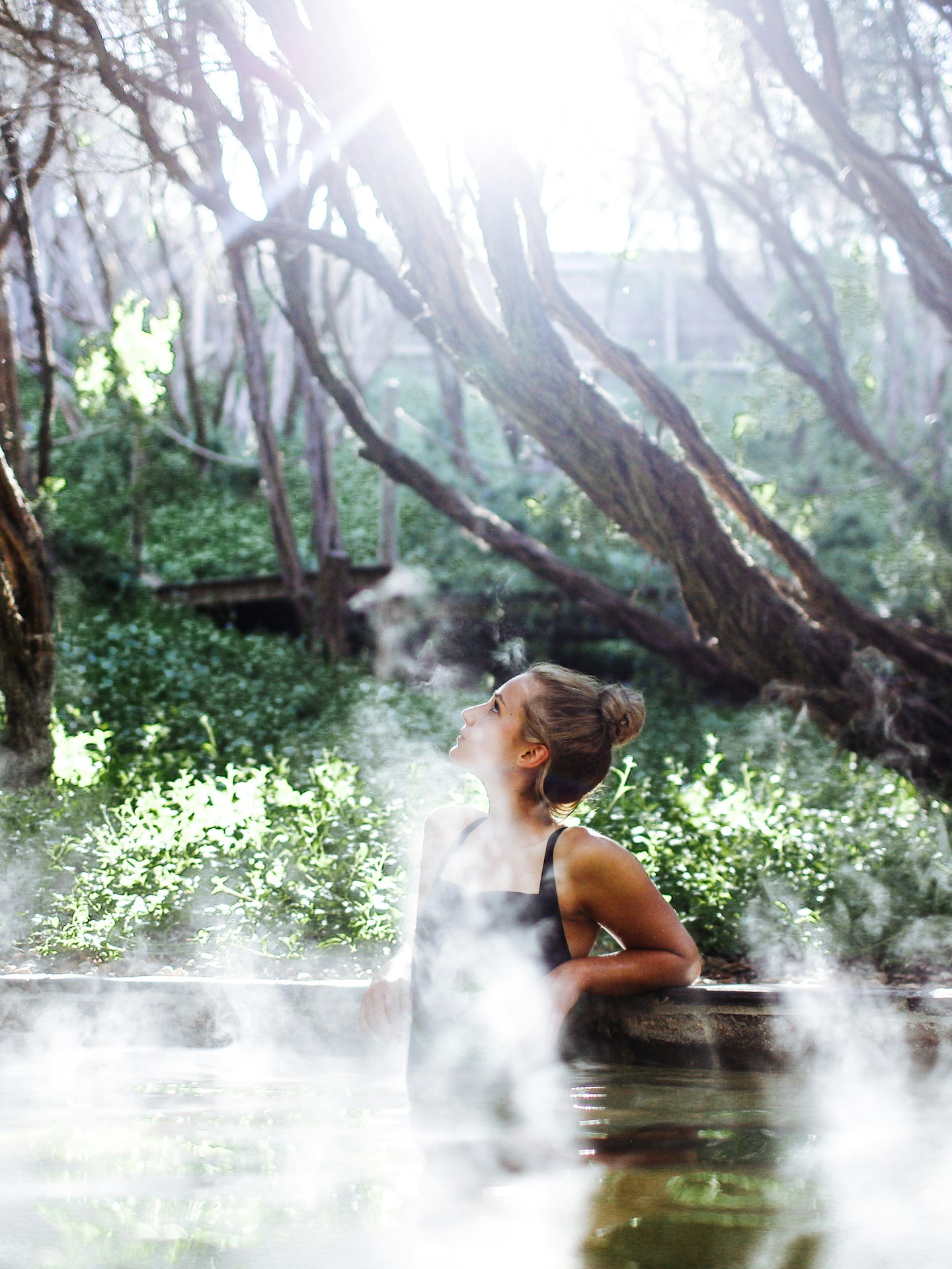woman in black bathing suit sitting in steamy geothermal pool looking up to the sky with nature all around