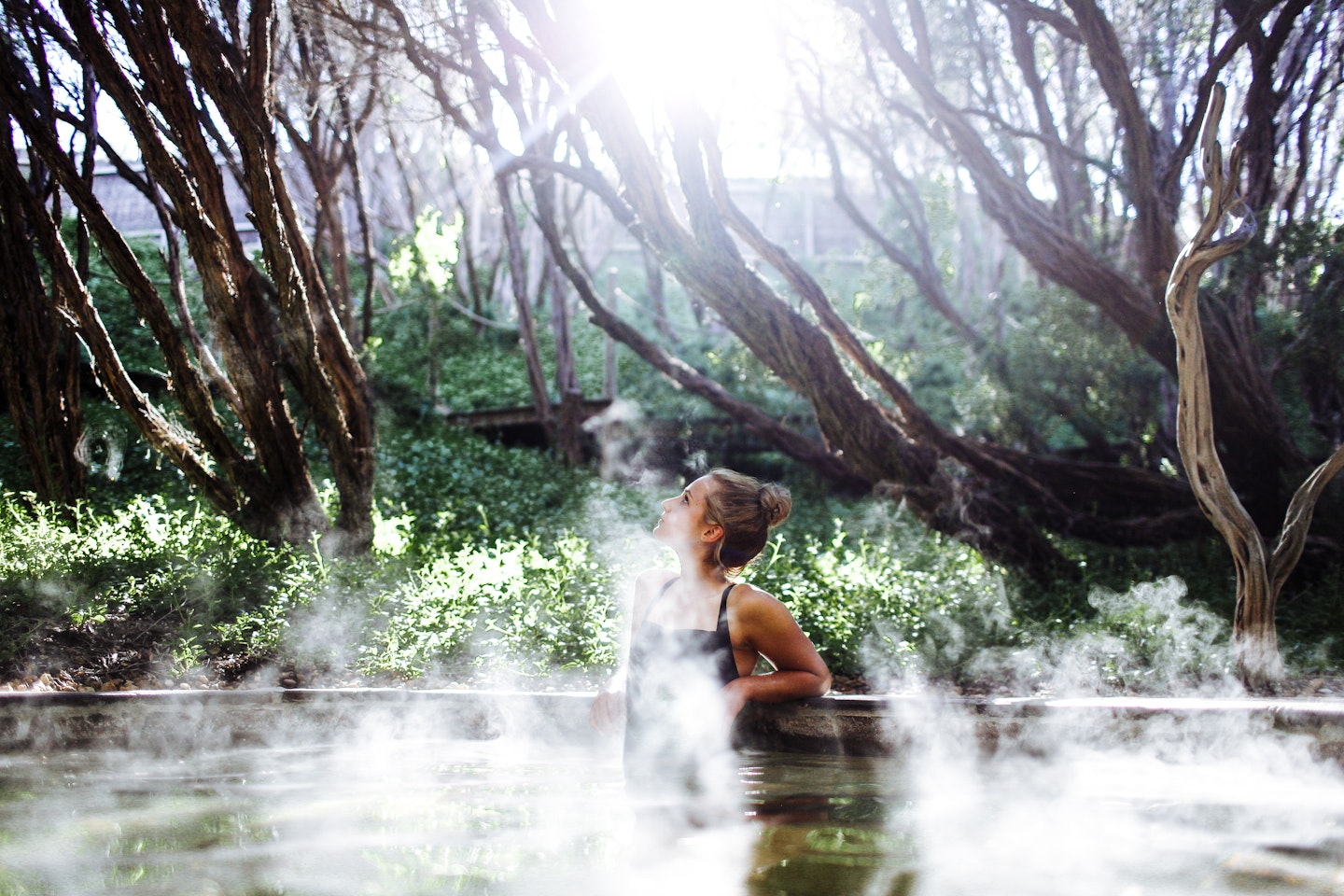 woman in black bathing suit sitting in steamy geothermal pool looking up to the sky with nature all around