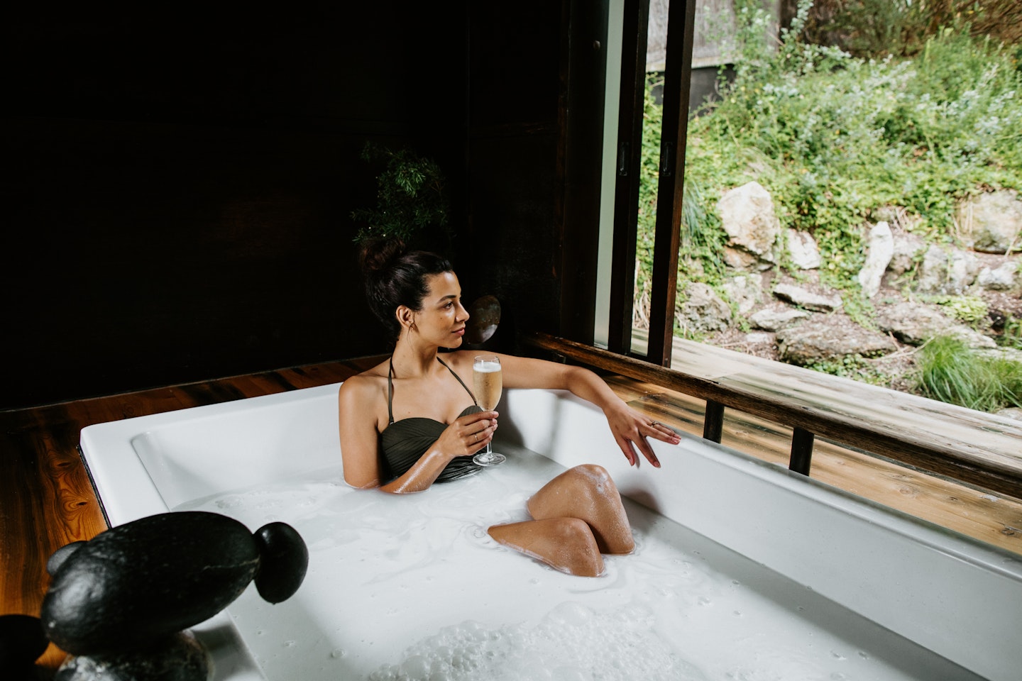 woman in bathing suit sitting in private bath with glass of champagne looking out at nature