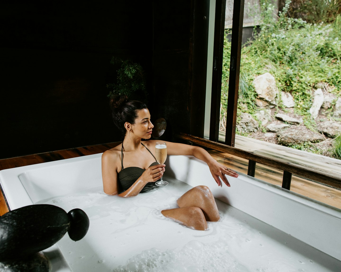 woman in bathing suit sitting in private bath with glass of champagne looking out at nature