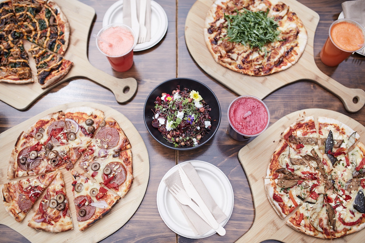 cafe table laden with pizzas on wooden boards, fresh salad and cold pressed juices