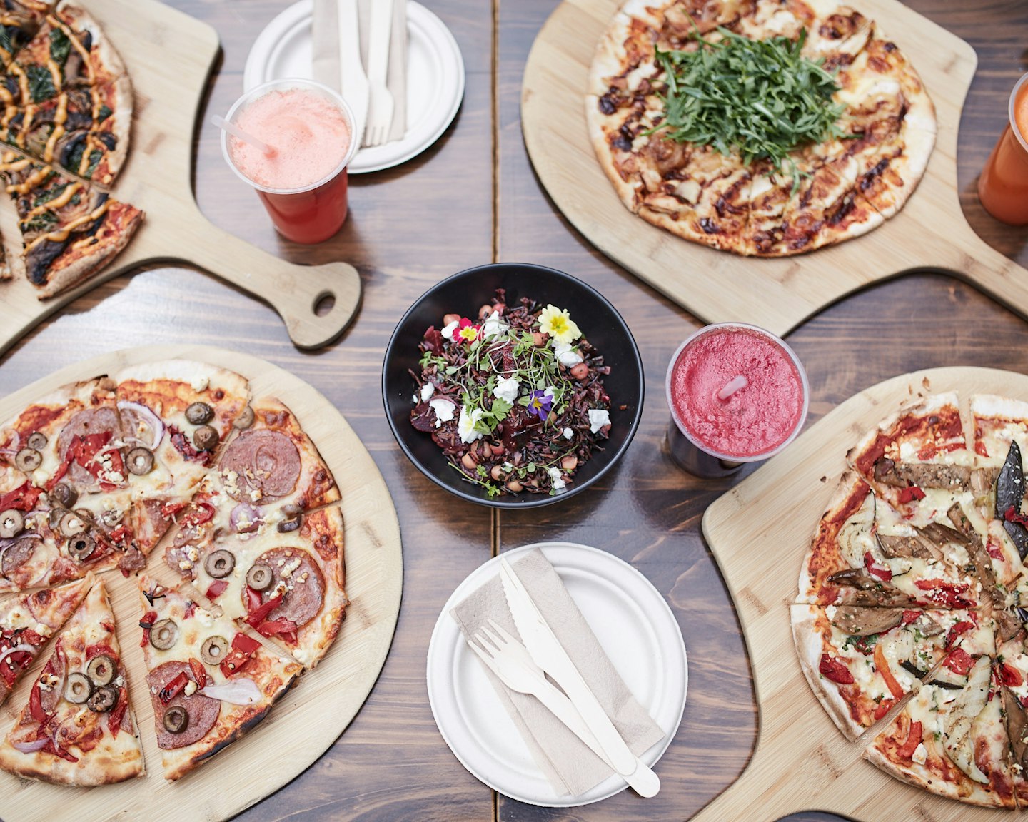cafe table laden with pizzas on wooden boards, fresh salad and cold pressed juices