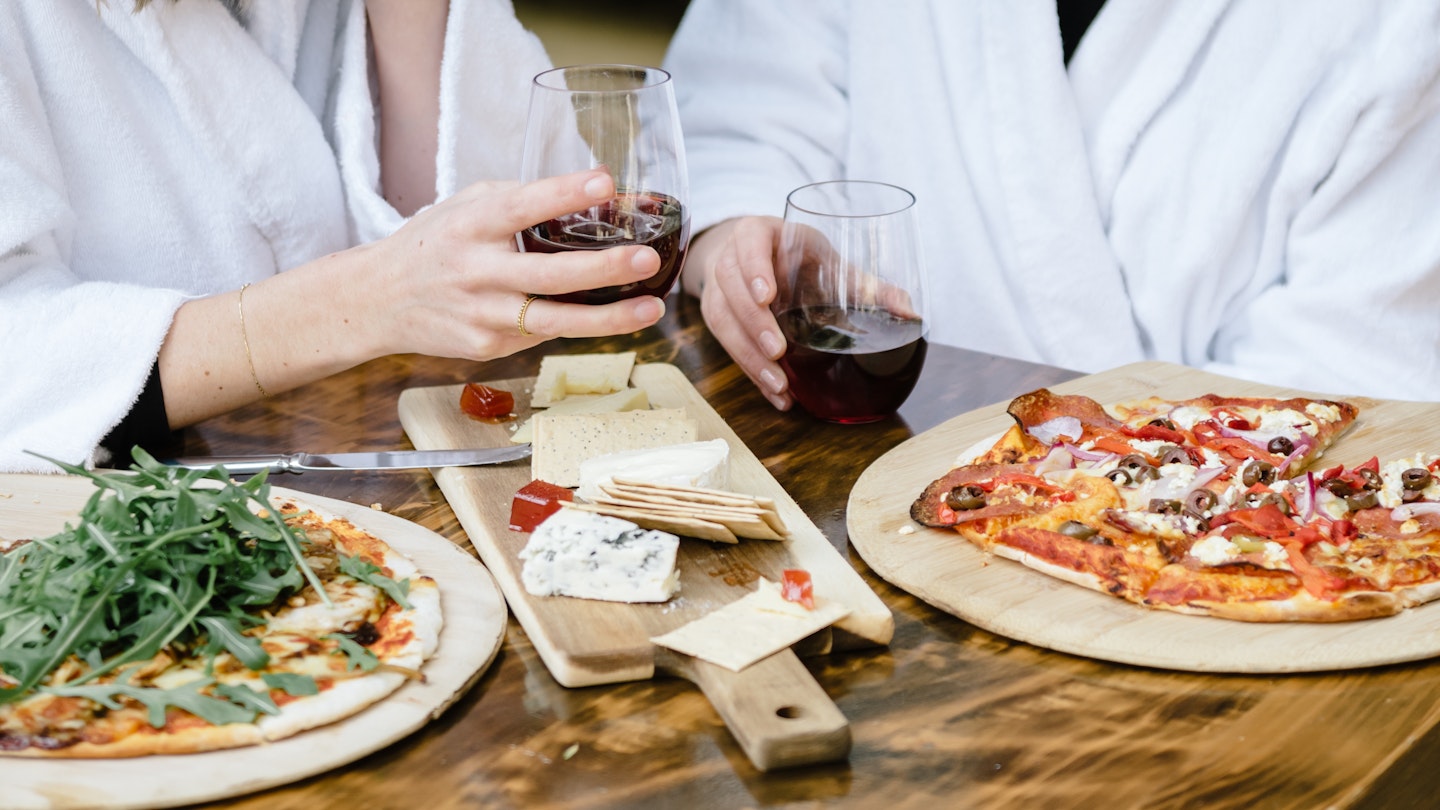 two people in white bath robes cheersing red wine glasses with pizzas and cheese board on table