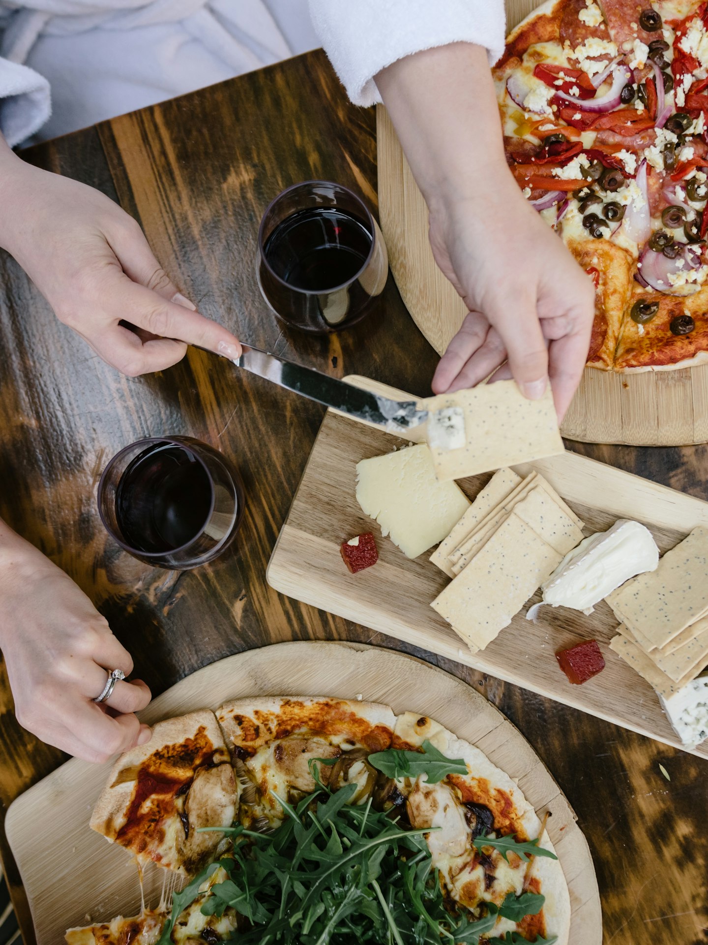 aerial view of two pizzas, a cheese board, two glasses of red wine on a café table and a person's hands and white bath robe 