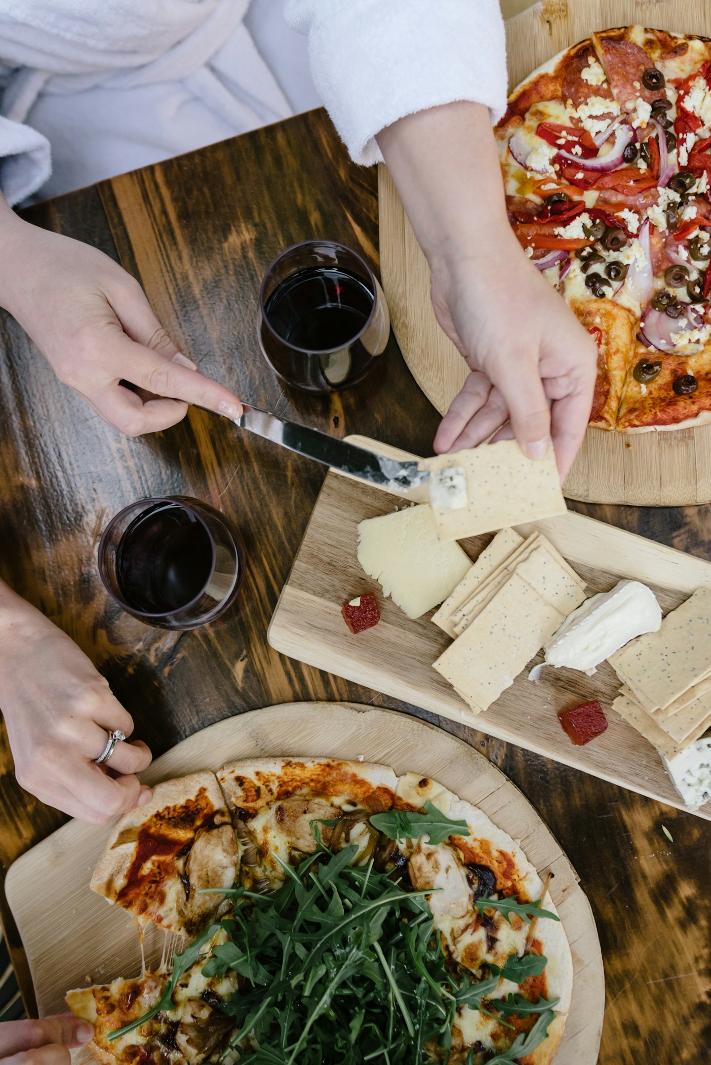 aerial view of two pizzas, a cheese board, two glasses of red wine on a café table and a person's hands and white bath robe 