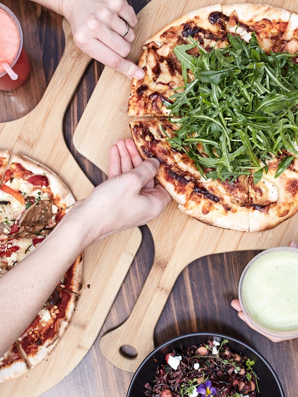 view from above of people's arms in white robes taking slices of pizza from boards with cold pressed juices and a salad in shot