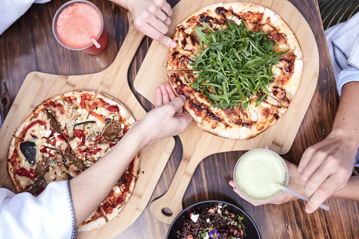 view from above of people's arms in white robes taking slices of pizza from boards with cold pressed juices and a salad in shot