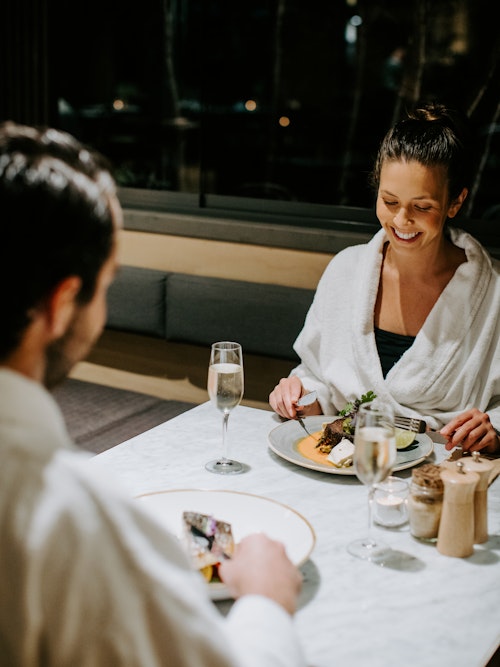 couple in white bath robes enjoying dinner in dining room with glasses of champagne