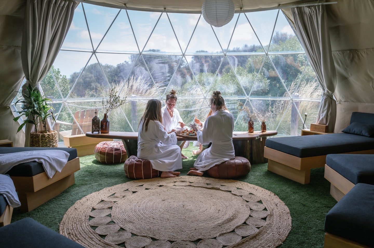 three women in white bath robes sitting on ottomans in dome space with daybeds and nature iew 