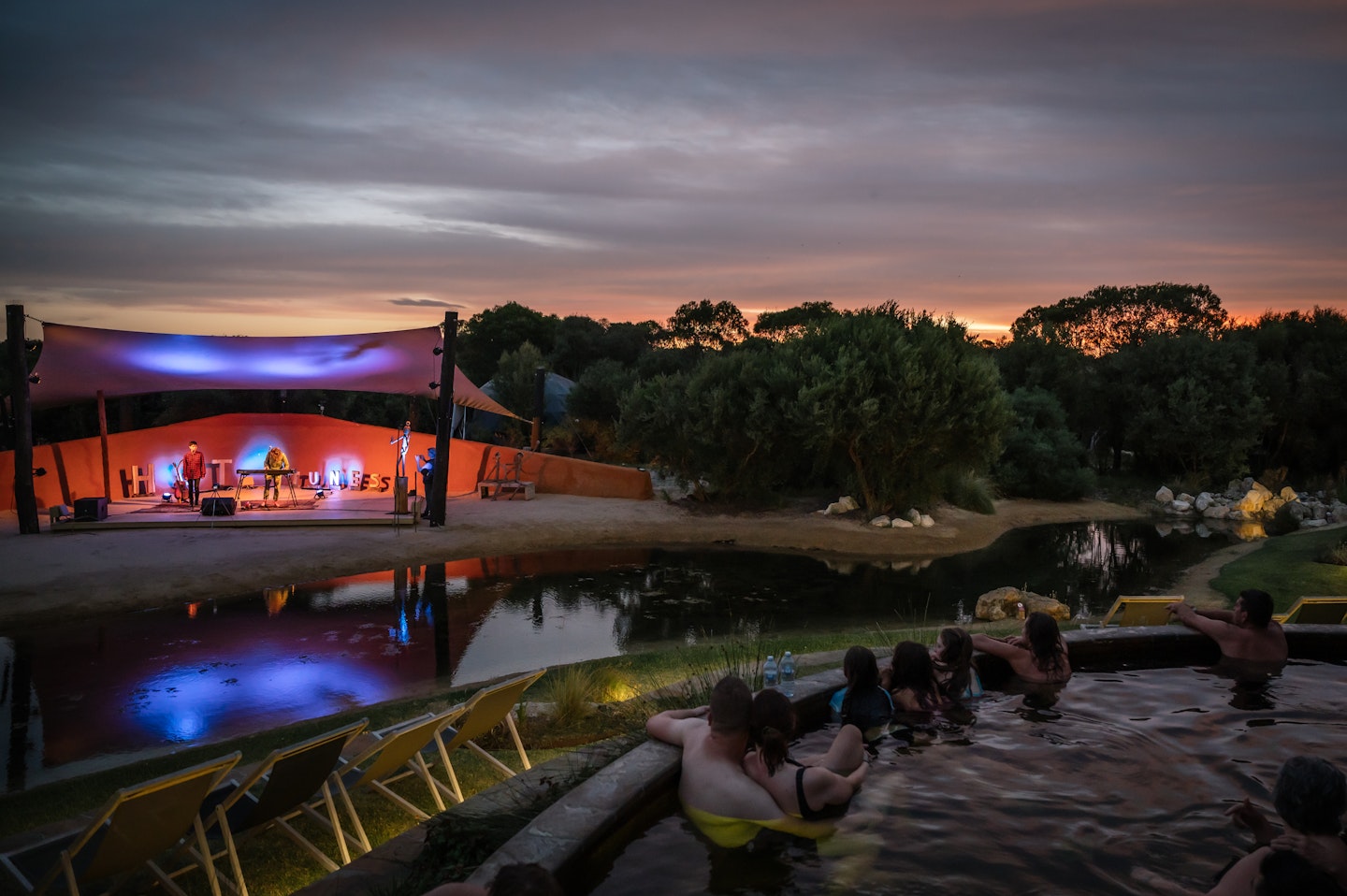 people sitting in amphitheatre hot pools with deck chairs on grass in front as they watch musical performance on lit up stage