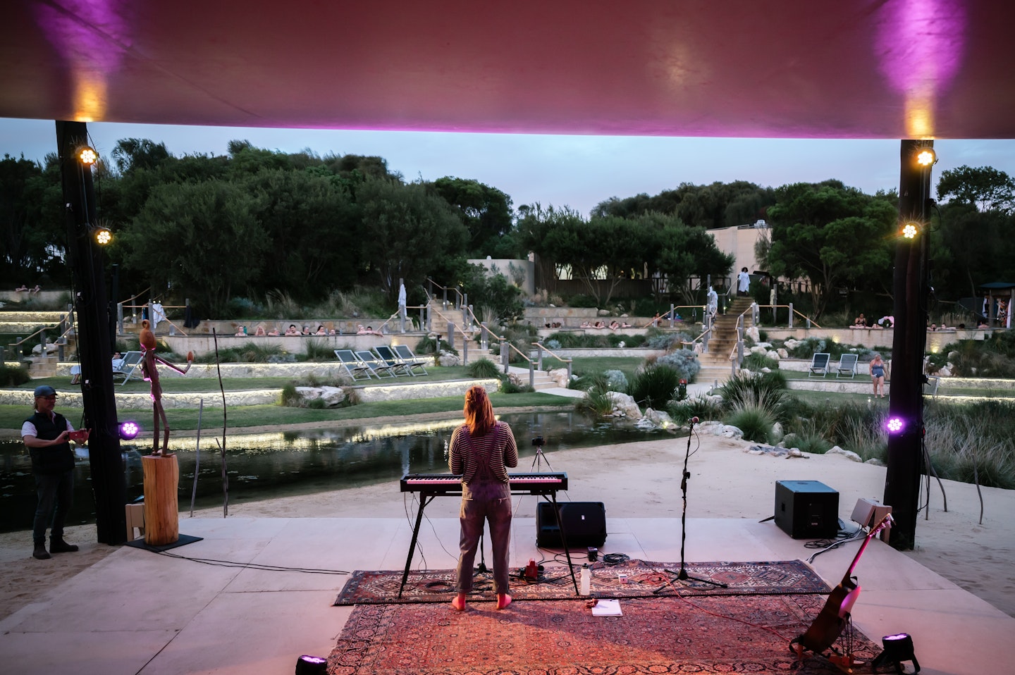 musician performing on amphitheatre stage looking out at pools with people and deck chairs