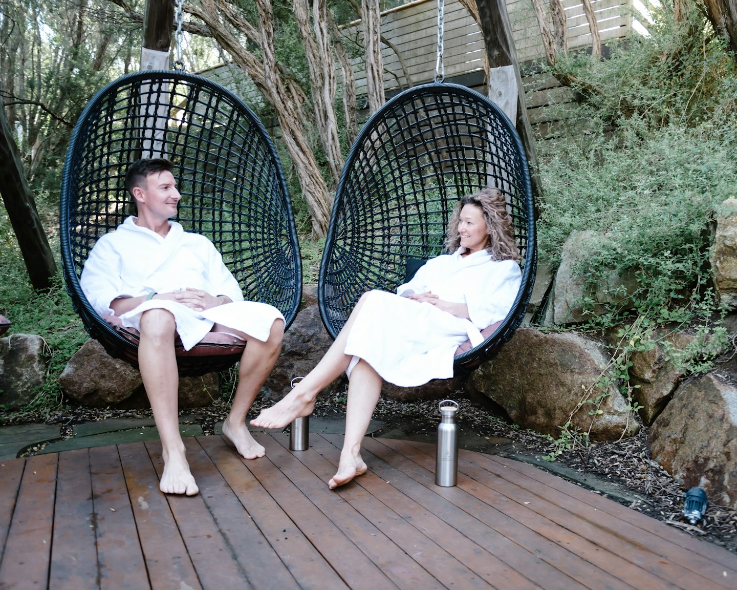 man and woman in white bath robes sit in hanging pod chairs in nature