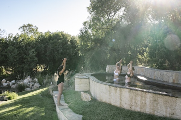 two women in bathing suits doing yoga in a hot pool with instructor standing on ledge in front of them