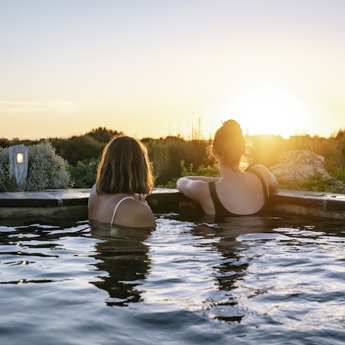 two young ladies in bathers sitting in geothermal hot pool looking out at view of nature and sunset
