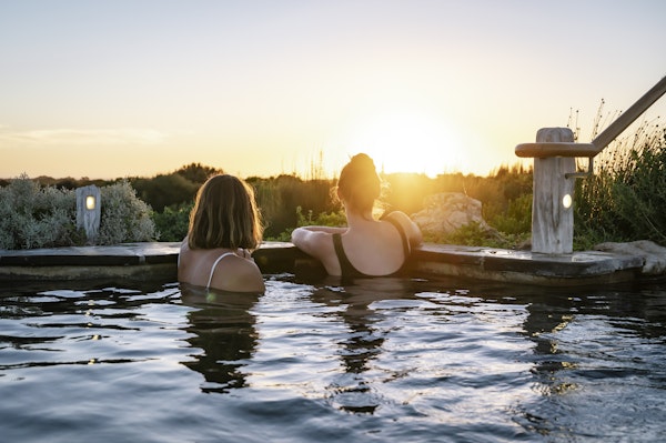 two young ladies in bathers sitting in geothermal hot pool looking out at view of nature and sunset