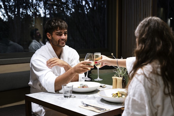 couple in white bath robes sitting at dining table clinking wine glasses