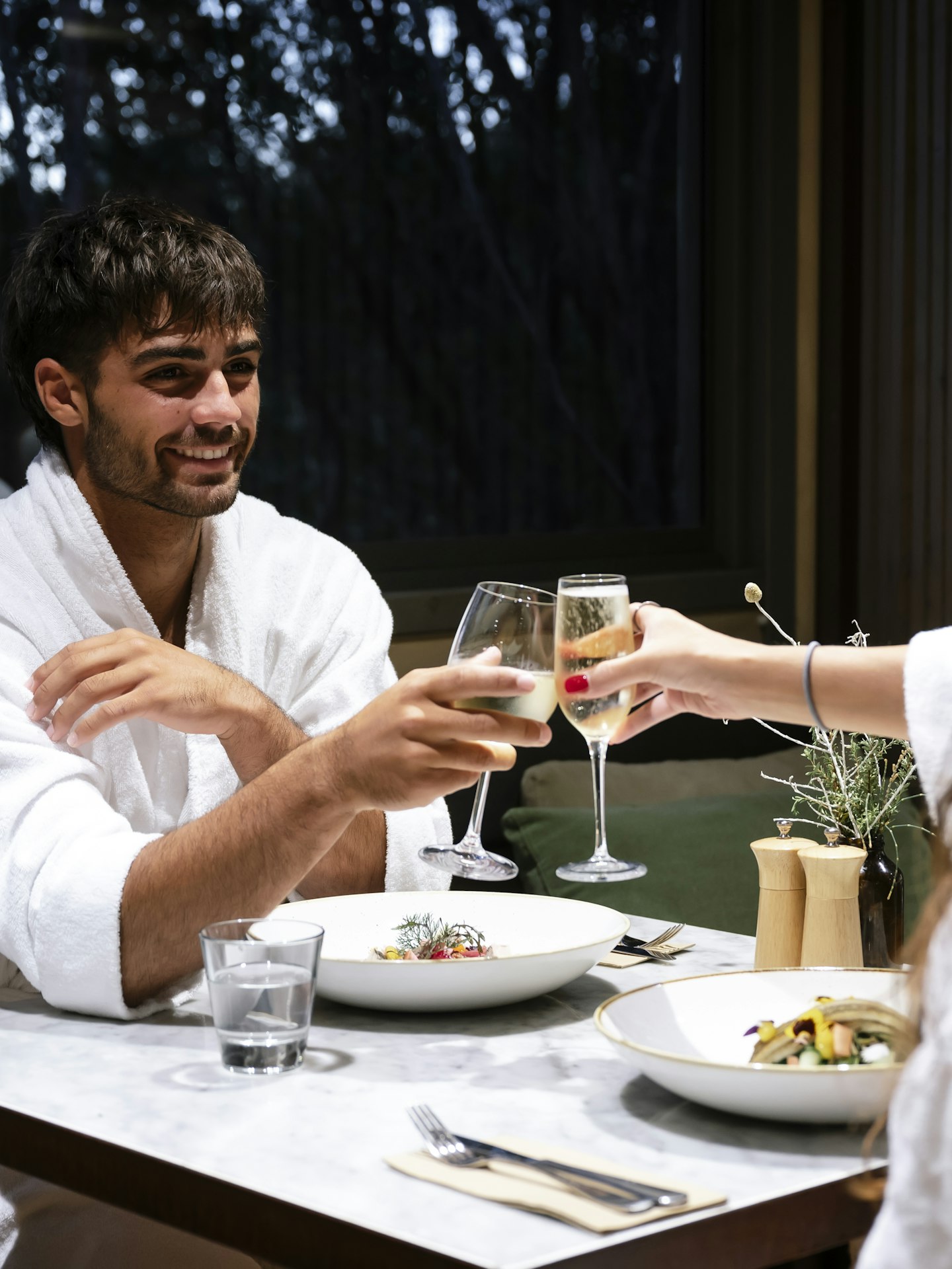 couple in white bath robes sitting at dining table clinking wine glasses