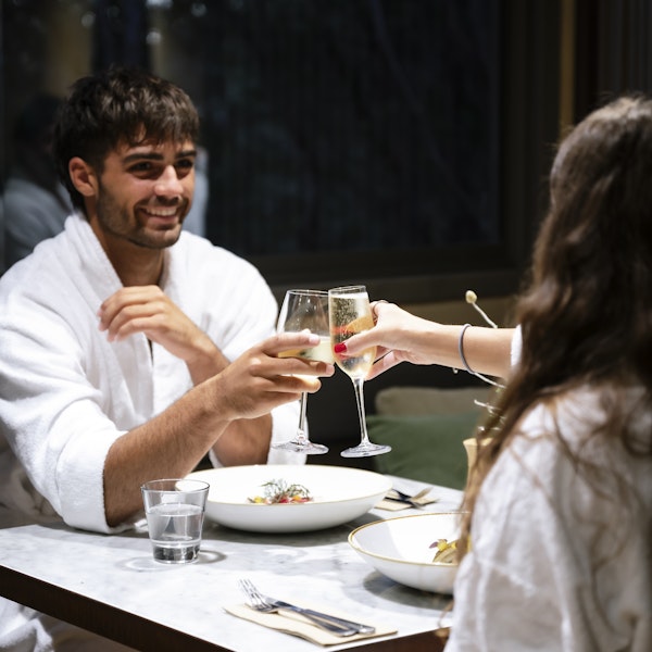 man in white bath robe sitting opposite woman in white bath robe clinking champagne glasses over dinner