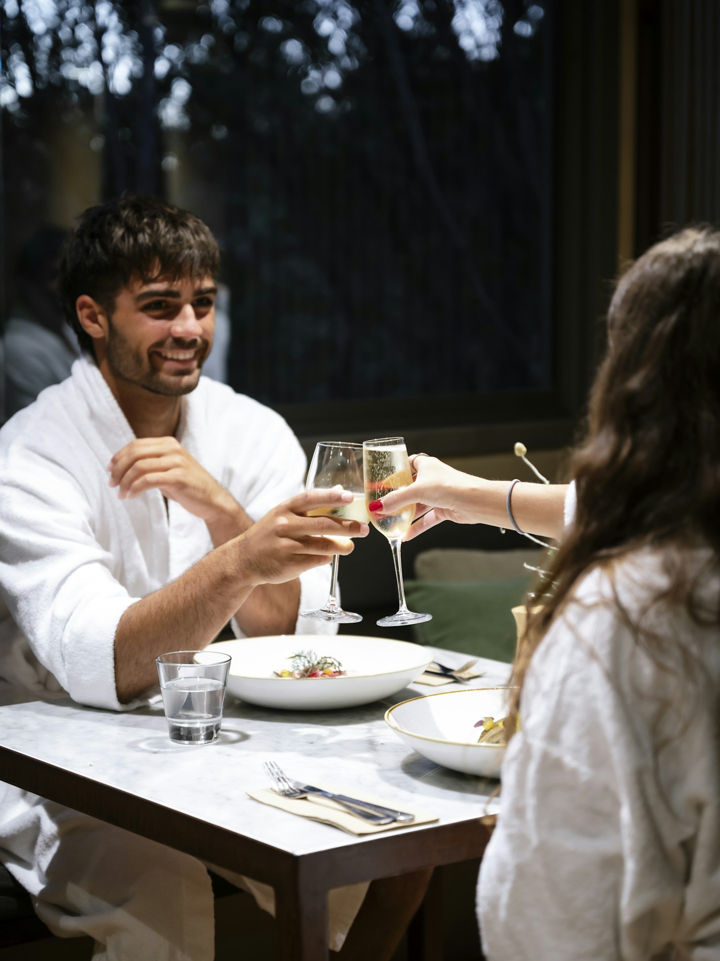 man in white bath robe sitting opposite woman in white bath robe clinking champagne glasses over dinner