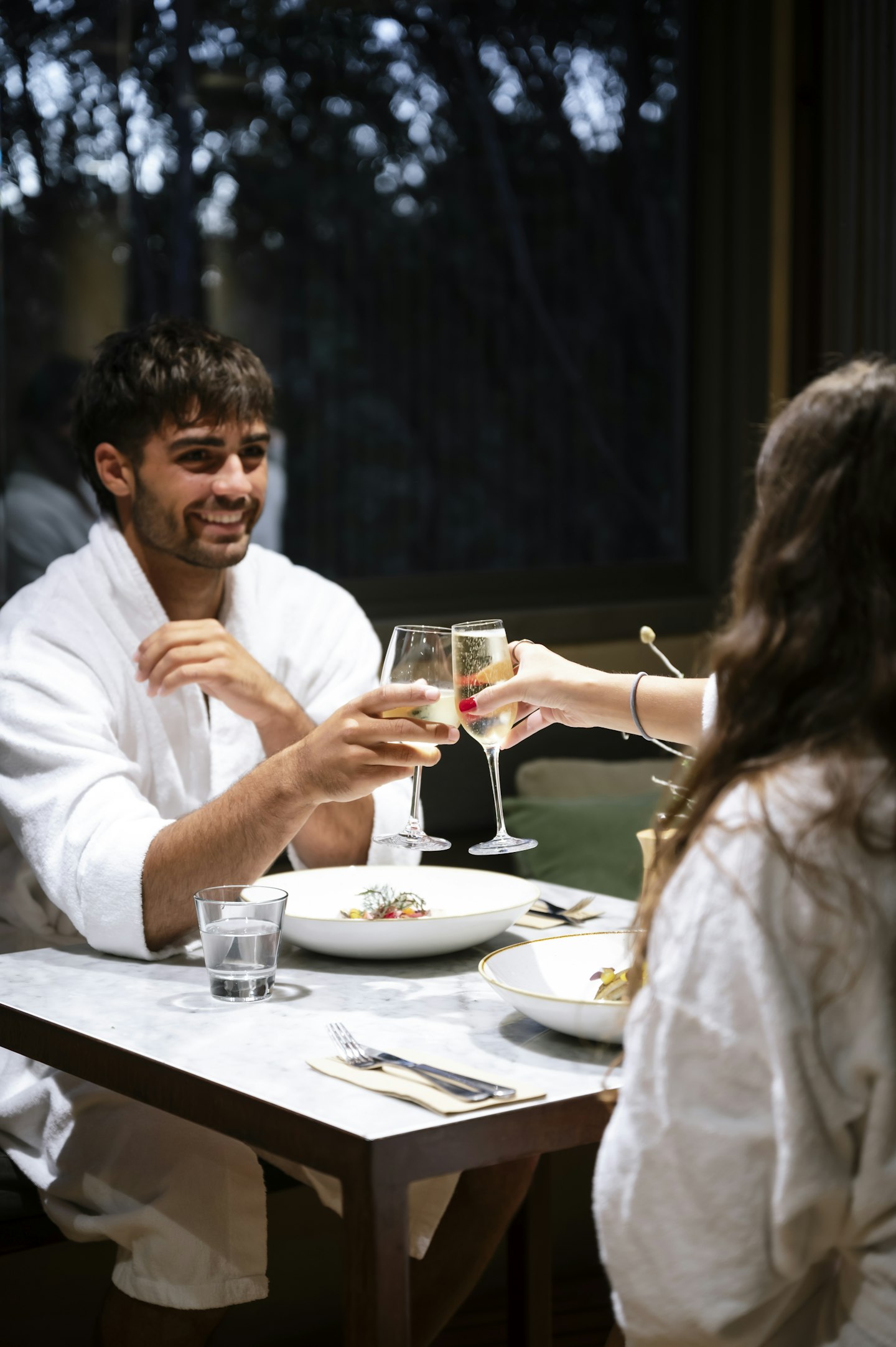 man in white bath robe sitting opposite woman in white bath robe clinking champagne glasses over dinner