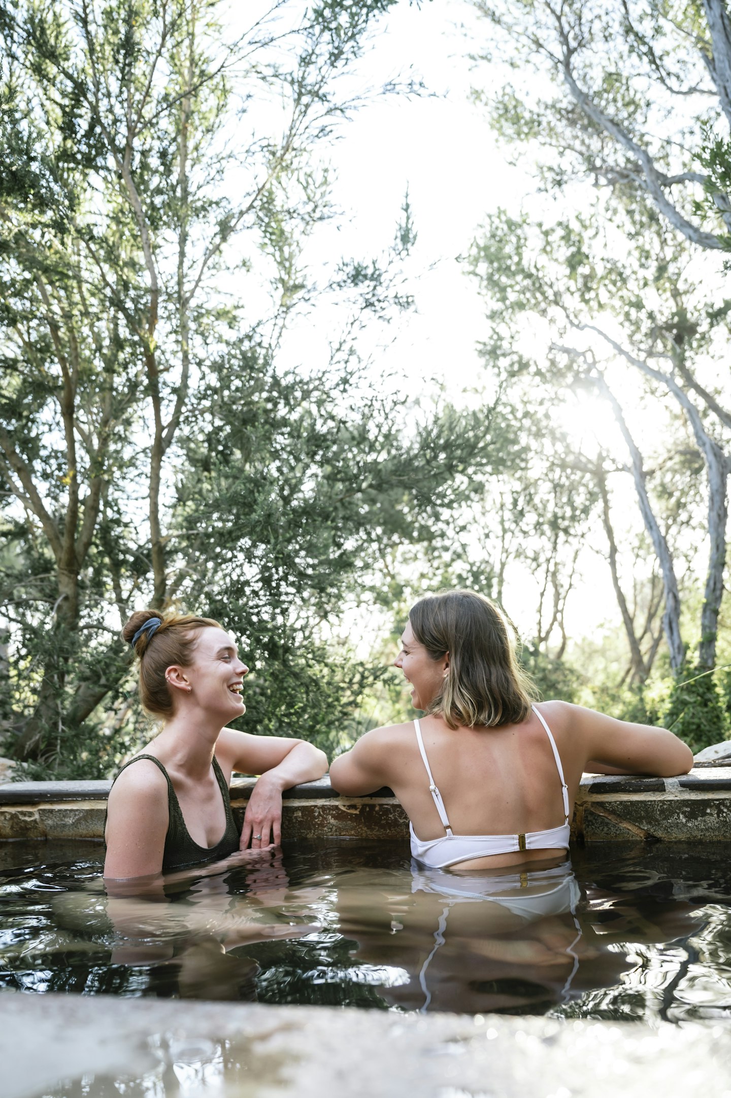 two young ladies in bathing suits laughing and relaxing in geothermal pool with sunlight breaking through trees