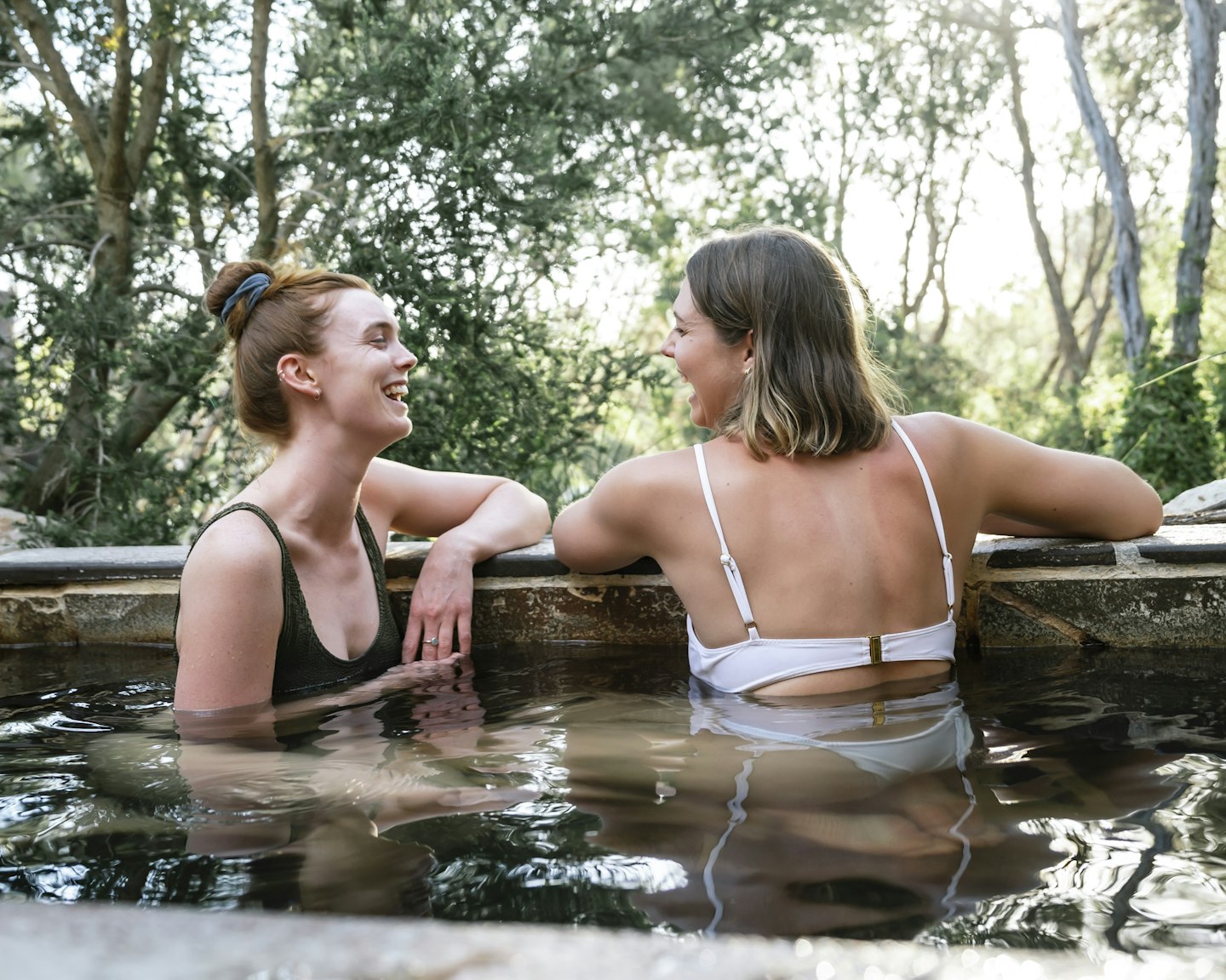 two young ladies in bathing suits laughing and relaxing in geothermal pool with sunlight breaking through trees