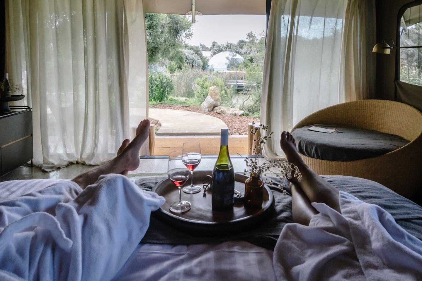 feet and legs of two people in white bath robes lying on bed looking out at nature view with wine and wine glasses on tray