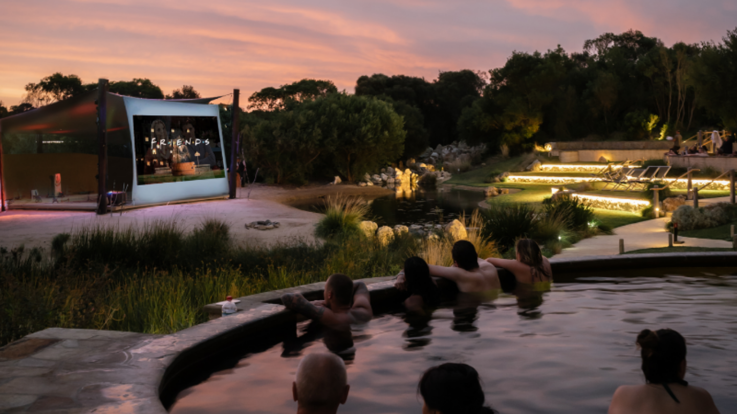 people sitting in geothermal pools watching FRIENDS on giant outdoor cinema screen at night