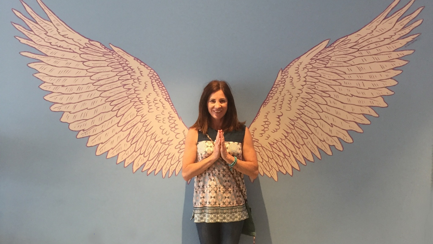 woman standing in front of wall with angel wings painted on it, her hands in prayer pose