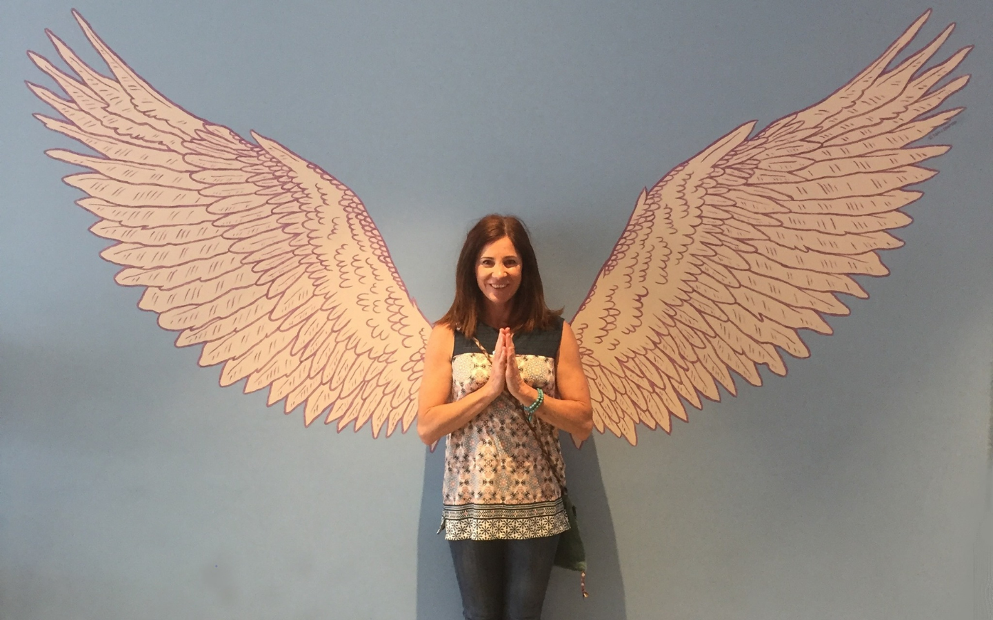 woman standing in front of wall with angel wings painted on it, her hands in prayer pose