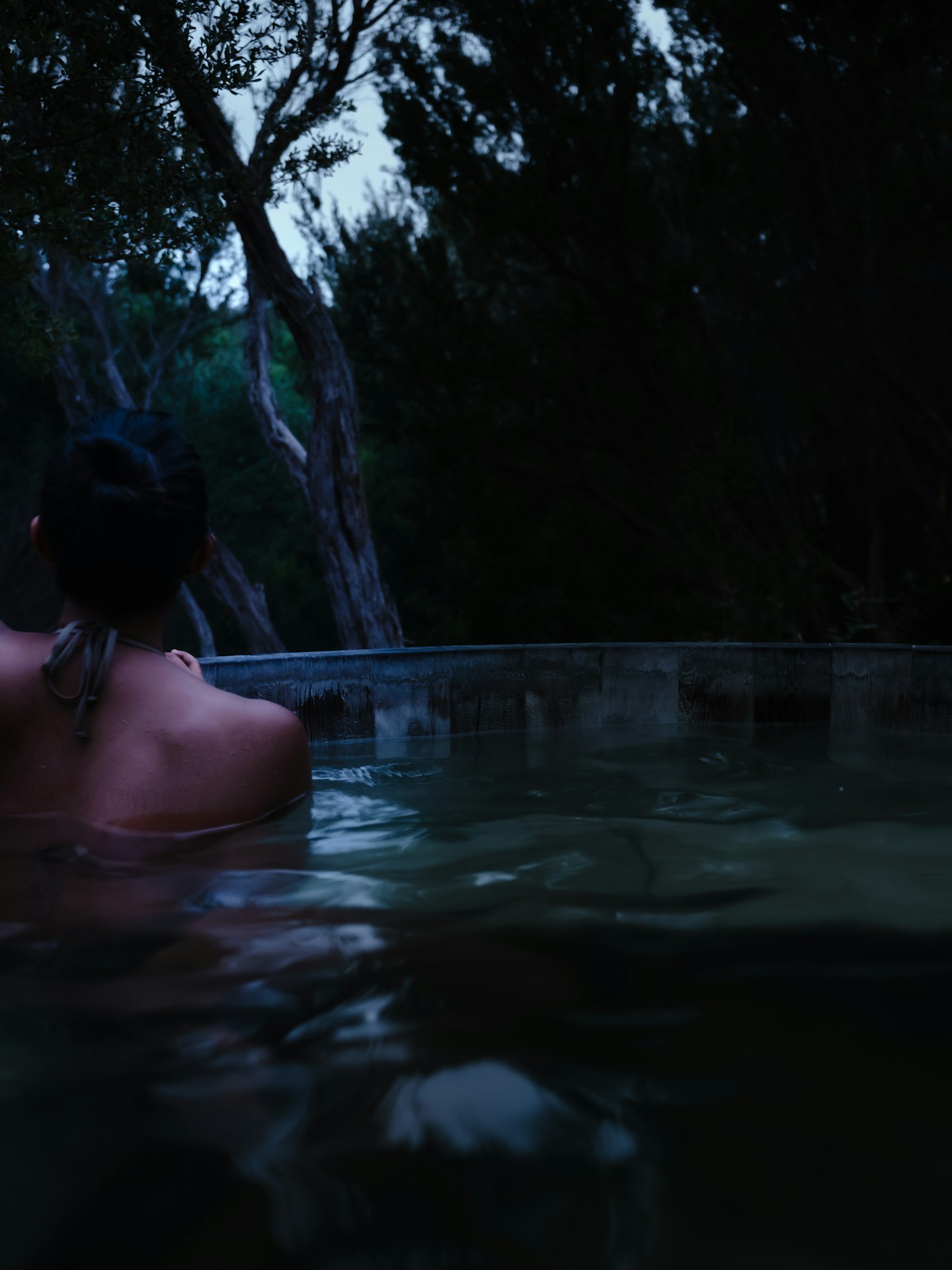 back of woman in bathing suit sitting in hot pool at night time