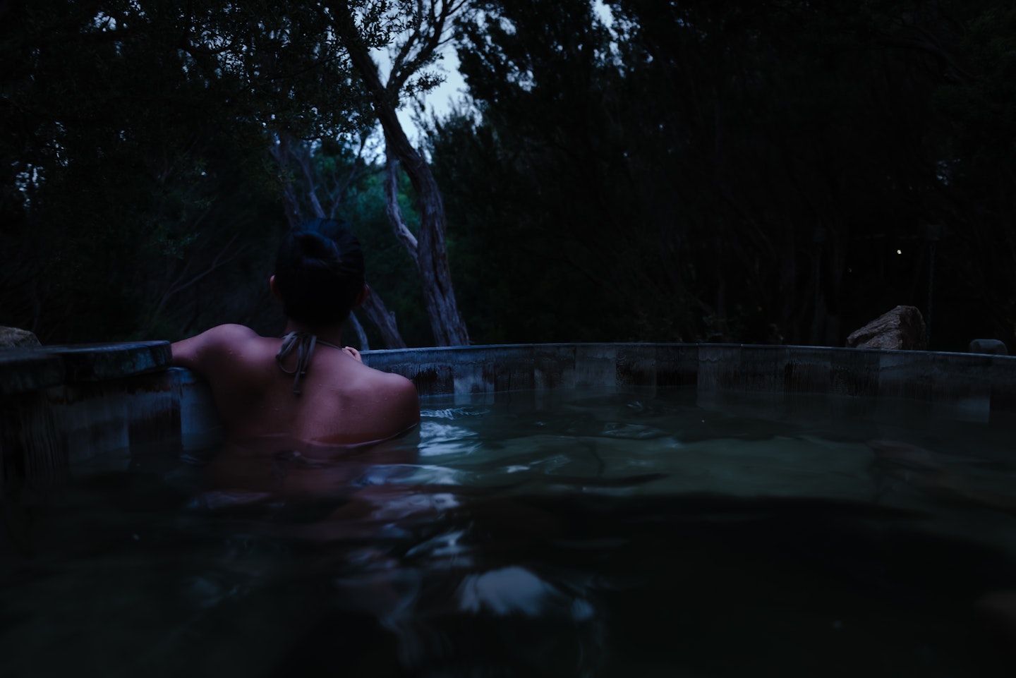 back of woman in bathing suit sitting in hot pool at night time