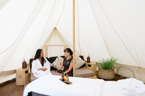 spa therapist and guest in white bath robe discuss treatment options in wellness consultation tent with massage table set up