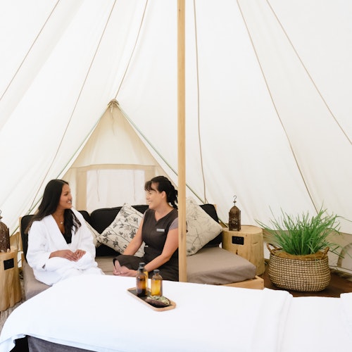 spa therapist and guest in white bath robe discuss treatment options in wellness consultation tent with massage table set up