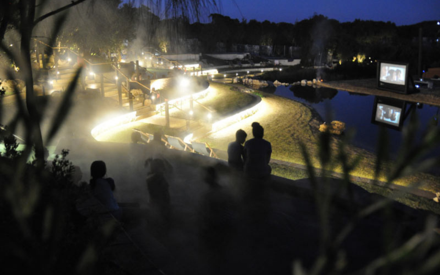 people sitting in geothermal pools at night time watching movie on giant outdoor cinema screen