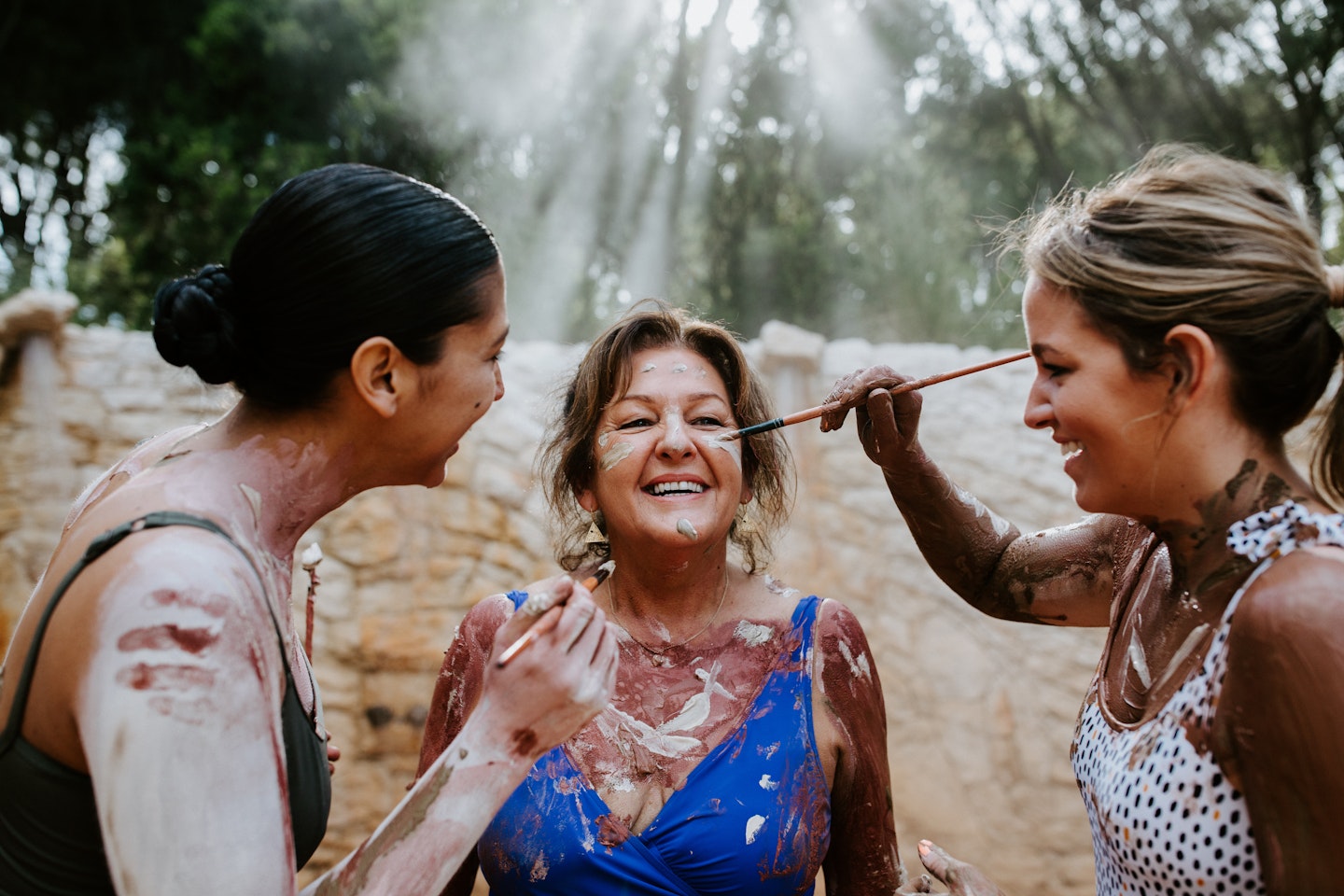 two women in bathing suits painting another woman in bathing suit's face with indigenous clay