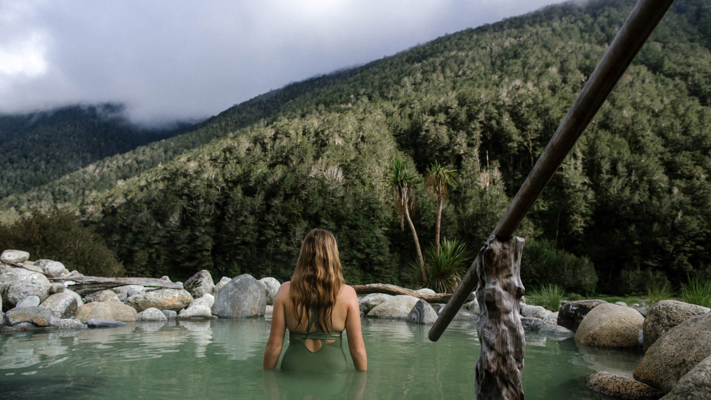 woman in bathing suit standing in geothermal pool looking out at mountain forrest