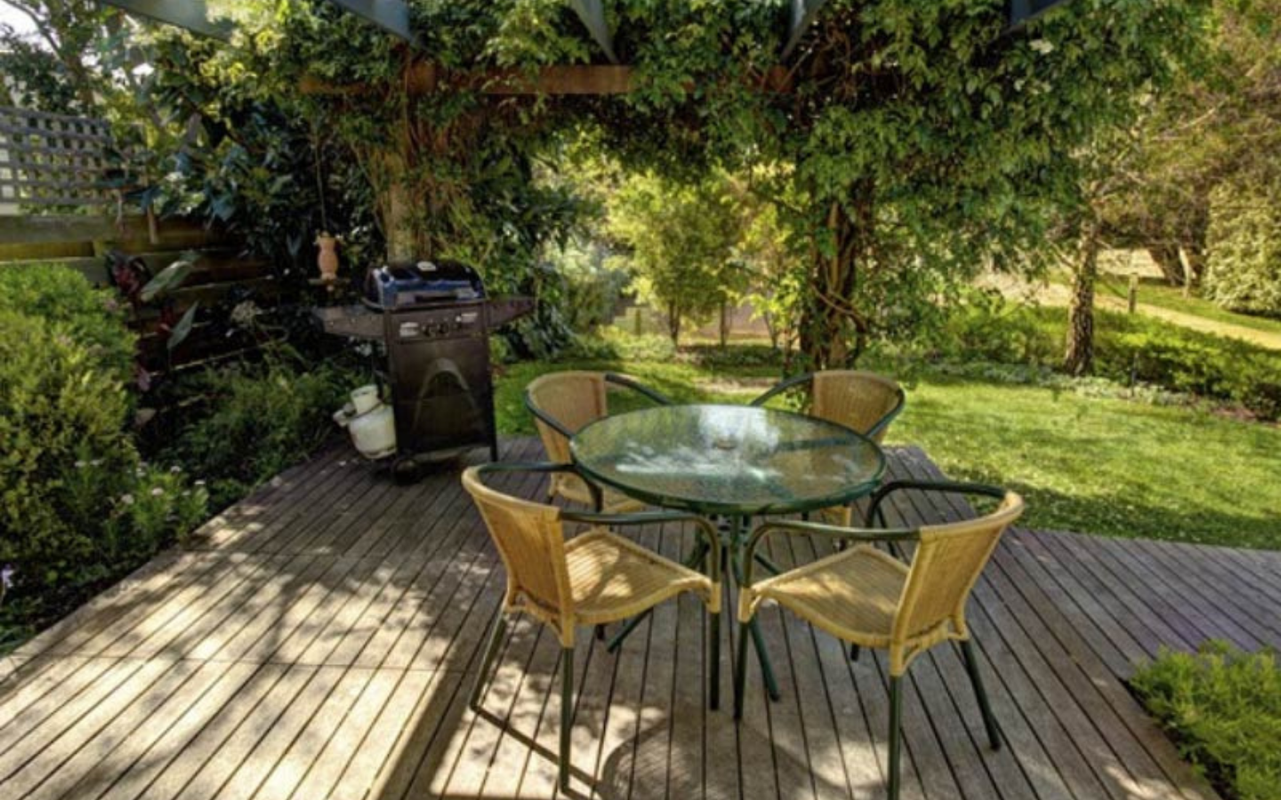 outdoor dining area with four chairs surrounding round glass table in front of a barbeque.