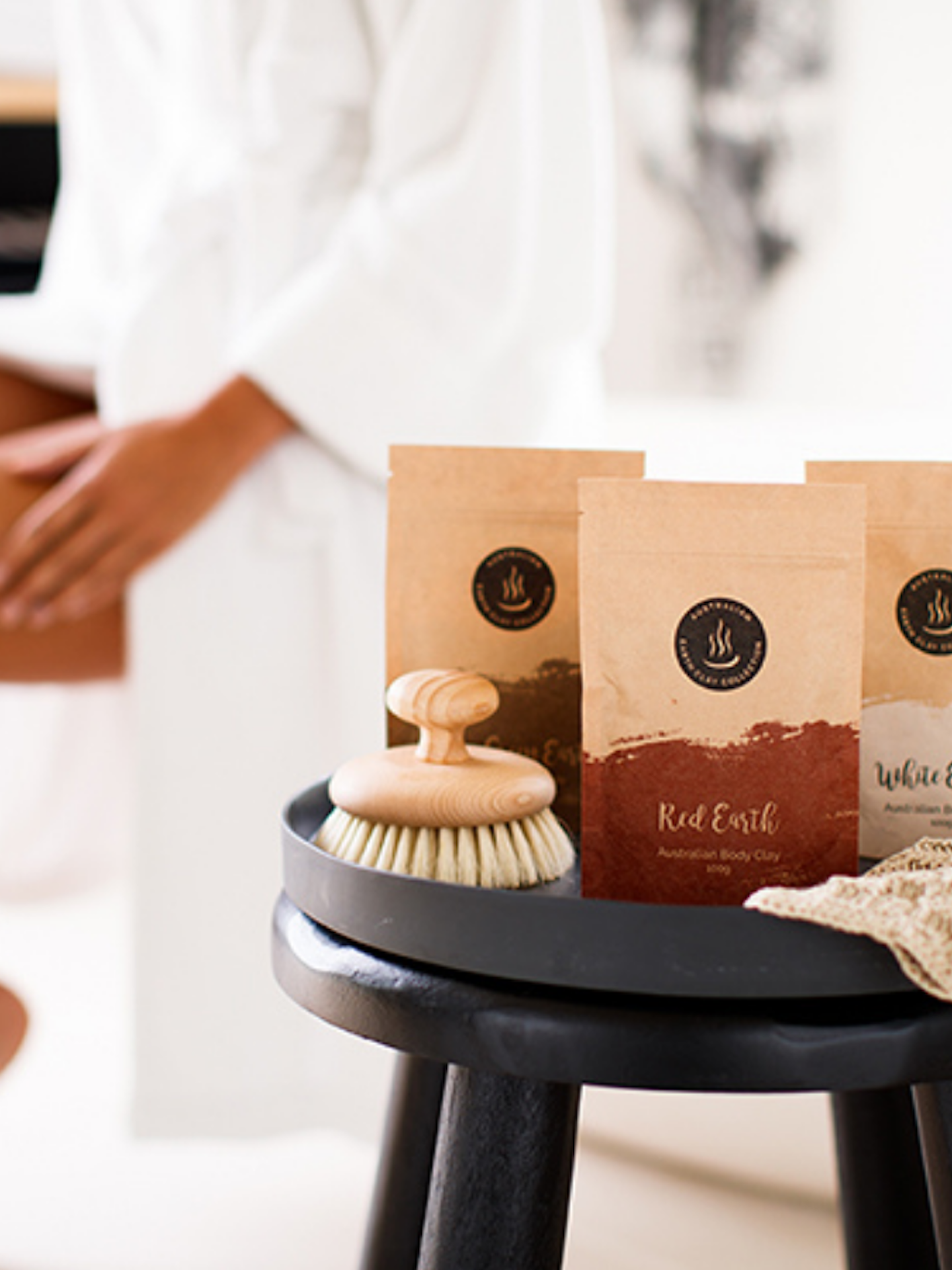 three bags of body clay on a stool with a woman sitting on a bath in a robe in the background