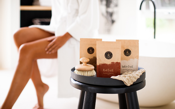 three bags of body clay on a stool with a woman sitting on a bath in a robe in the background