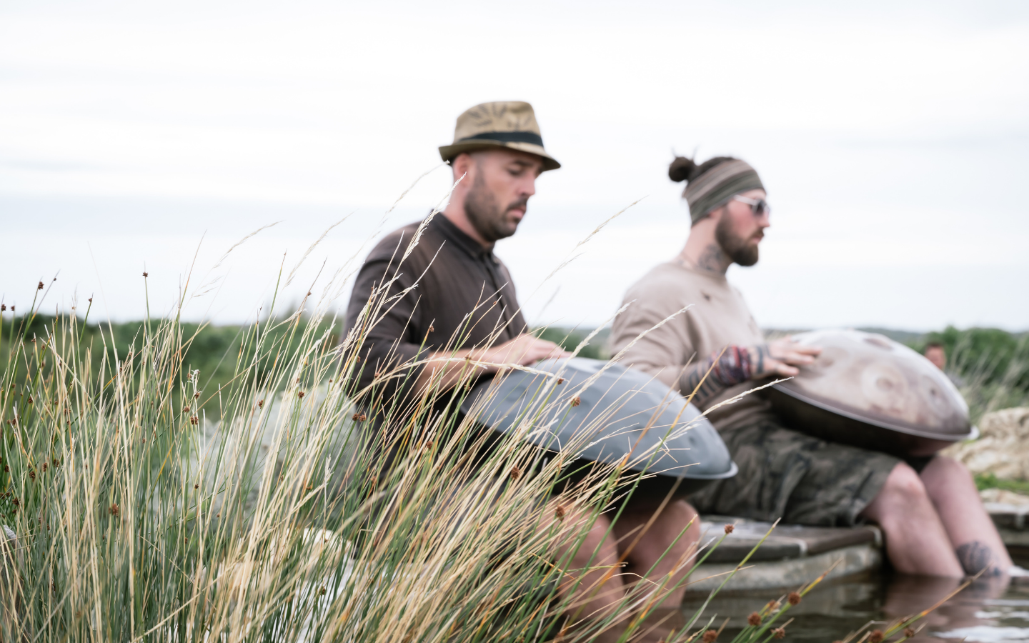 two men sitting in nature with feet in geothermal hot pool playing hand pans