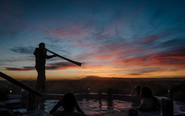 man playing yidaki at edge of hilltop hot pool at sunrise