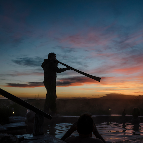 man playing yidaki at edge of hilltop hot pool at sunrise