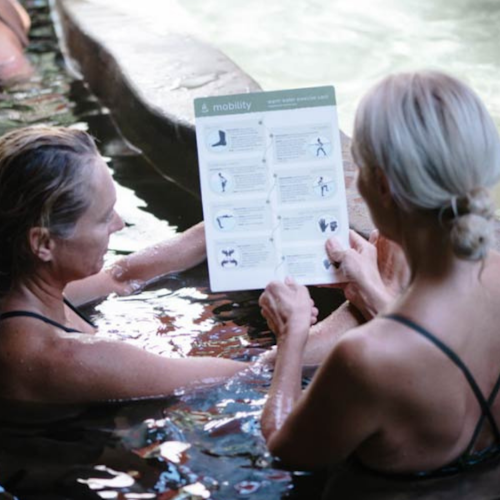 two women in bathing suits in hydrotherapy pool holding and reading warm water exercise instruction card