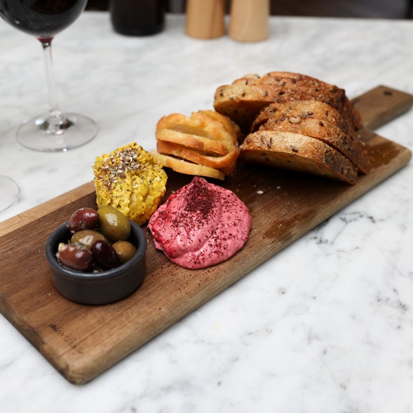 wooden board with dish of olives, two mounds of dips, a selection of toasted artisan breads