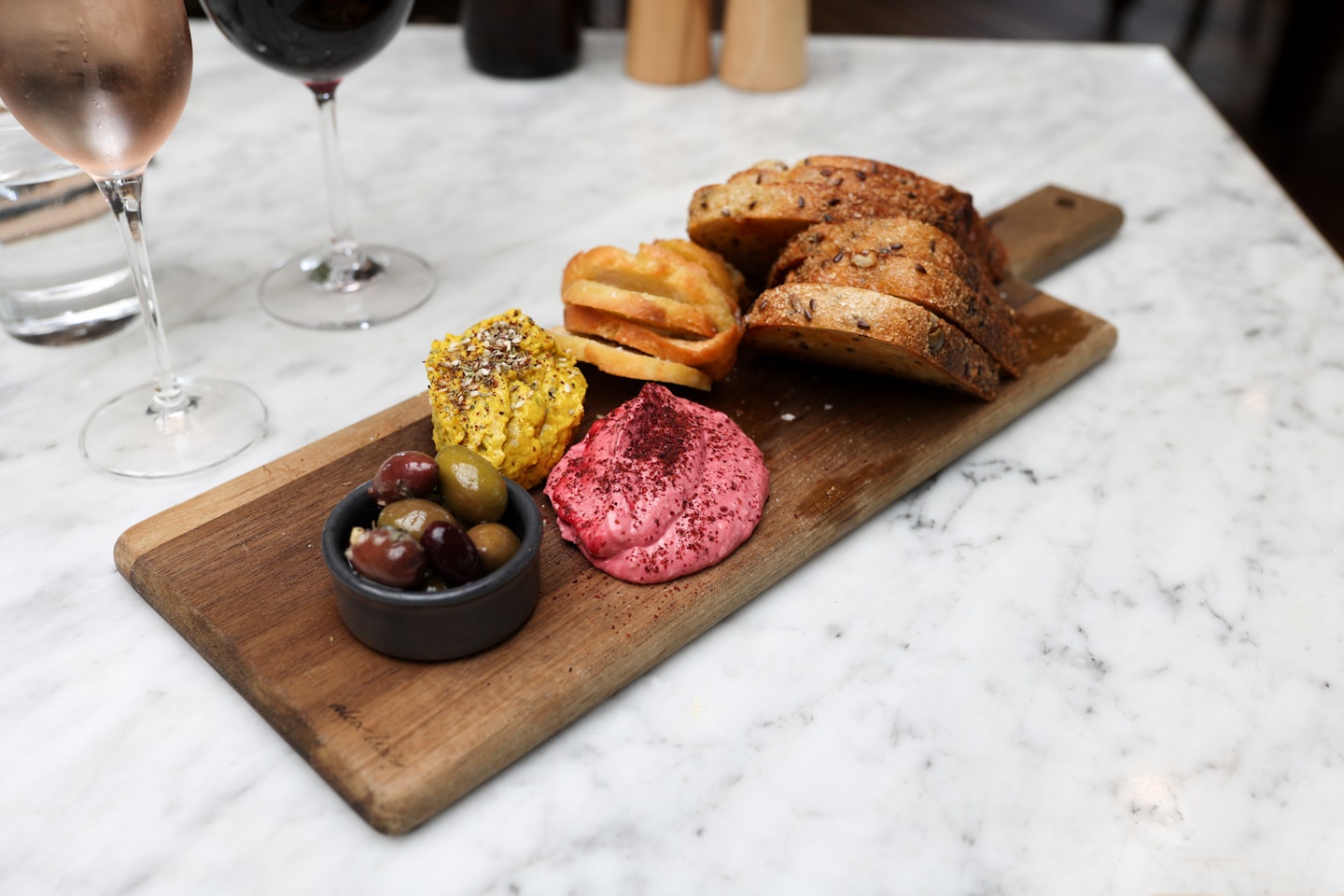 wooden board with dish of olives, two mounds of dips, a selection of toasted artisan breads
