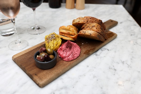 wooden board with dish of olives, two mounds of dips, a selection of toasted artisan breads