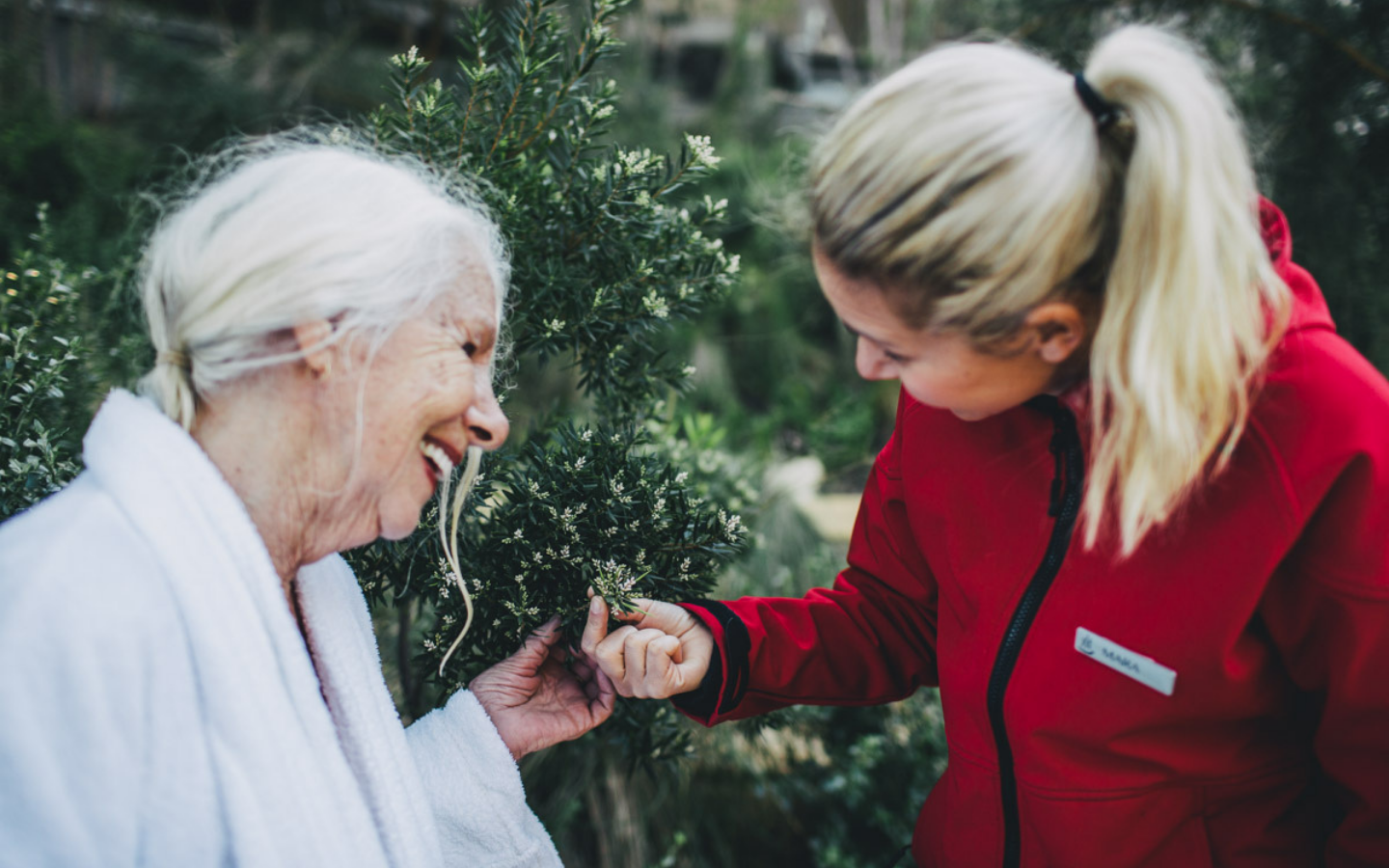 older lady in white bath robe and pool attendant in red jacket sharing a smile and holding foliage of a tree at the hot springs