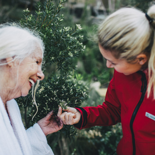 older lady in white bath robe and pool attendant in red jacket sharing a smile and holding foliage of a tree at the hot springs